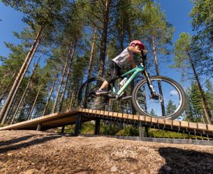 Barn med rosa hjälm cyklar på en teknikbana i skogen, på en grön mountainbike över en upphöjd träbro med tallar och blå himmel i bakgrunden.
