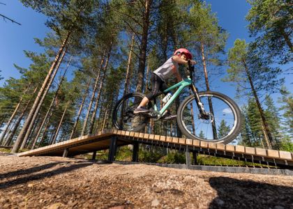 Barn med rosa hjälm cyklar på en teknikbana i skogen, på en grön mountainbike över en upphöjd träbro med tallar och blå himmel i bakgrunden.