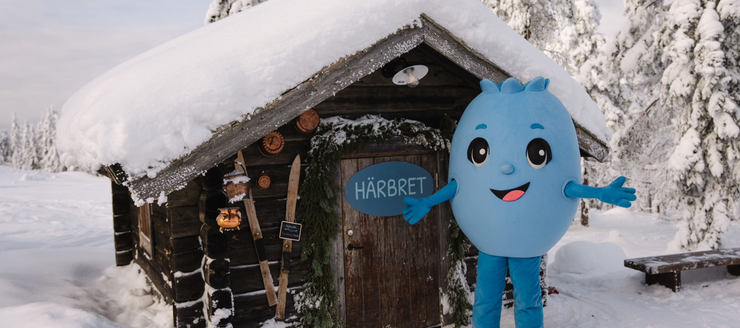 Blå figur står utanför en snötäckt timmerstuga med skylt på dörren i vintermiljö.