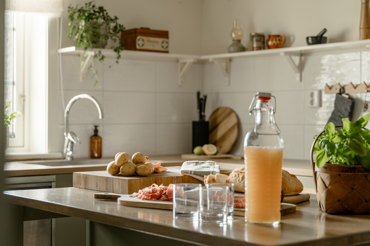 Kök med ljusa kakelväggar och öppna hyllor, där potatis, charkuterier, bröd, basilika och en flaska dryck står framme på köksön i förberett matsällskap.