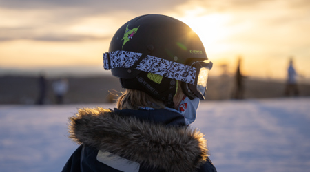 Person i skidhjälm och skidglasögon blickar ut över skidbacken i solnedgång.