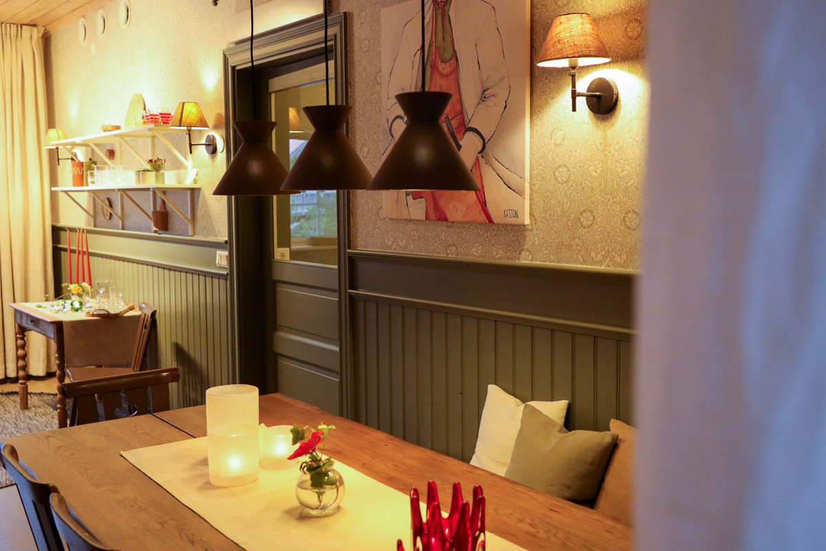 Cozy dining room with lit candles on a wooden table, pendant lights above, and artwork on the softly lit walls.