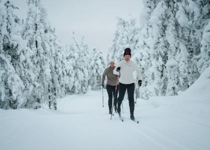 Två personer åker längdskidor på ett preparerat spår genom en snötäckt skog.