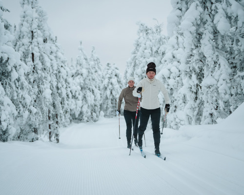 Two people cross-country skiing on a groomed trail through a snow-covered forest.
