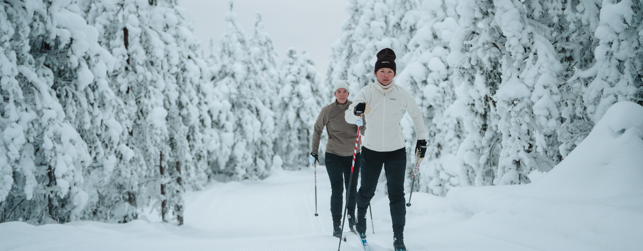Two people cross-country skiing on a groomed trail through a snow-covered forest.