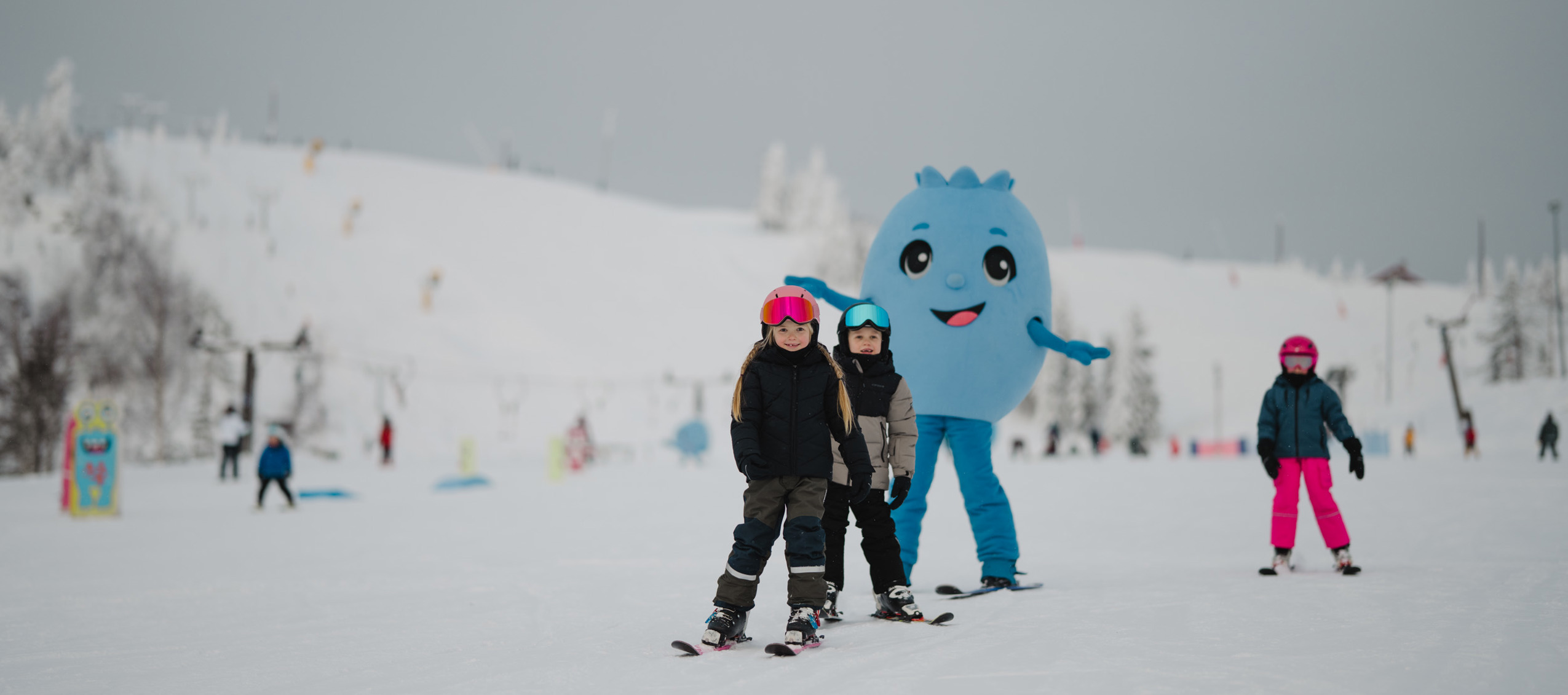 Barn åker skidor i en skidbacke tillsammans med en blå maskot i bakgrunden.