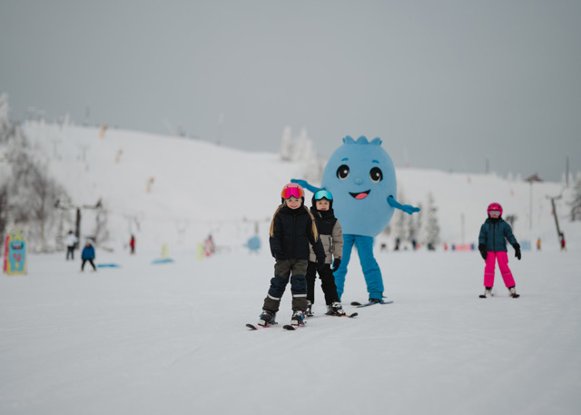 Barn åker skidor i en skidbacke tillsammans med en blå maskot i bakgrunden.