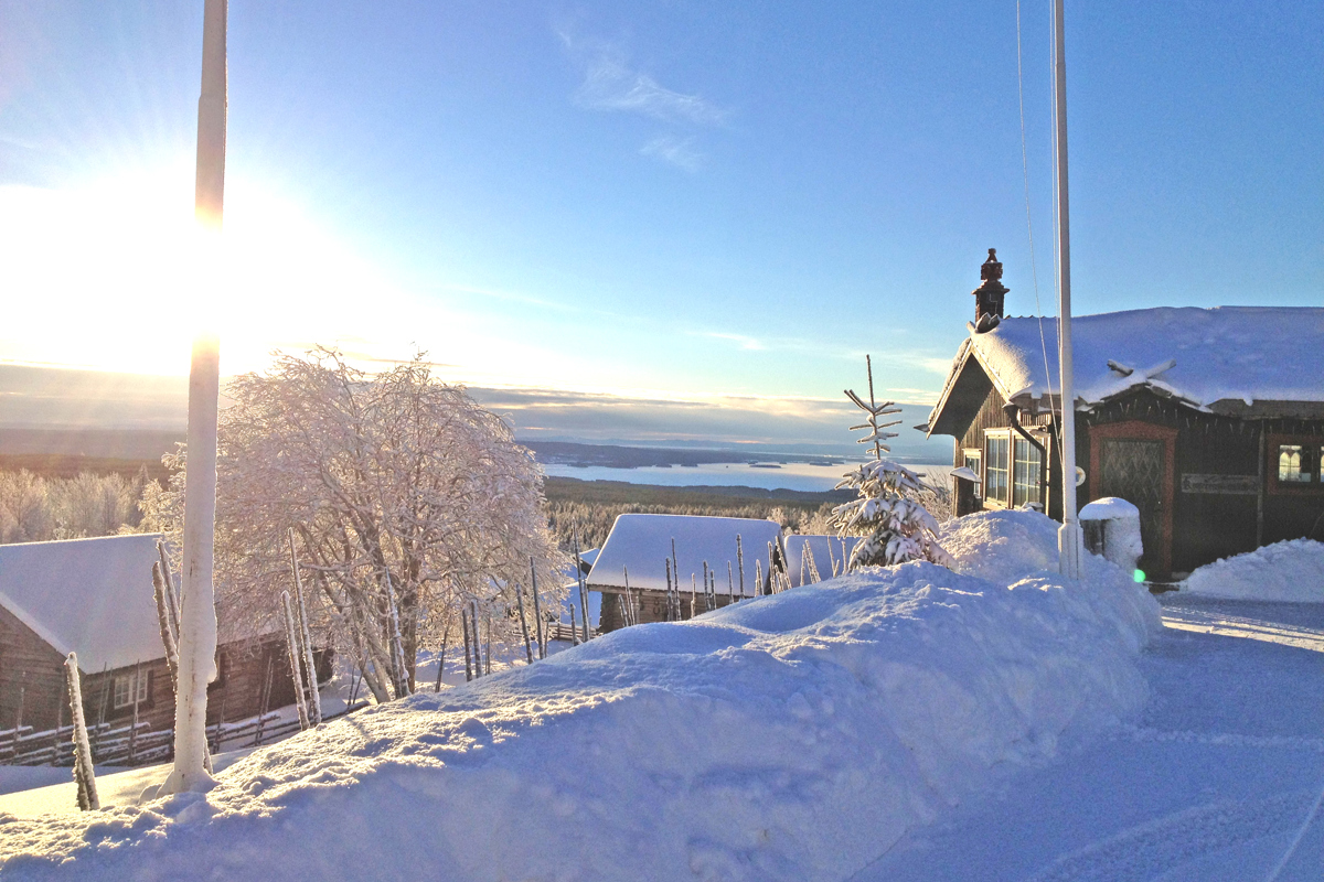 Winter view from Fryksås with snow-covered wooden houses, frosted trees, and the sun rising over Lake Orsa.