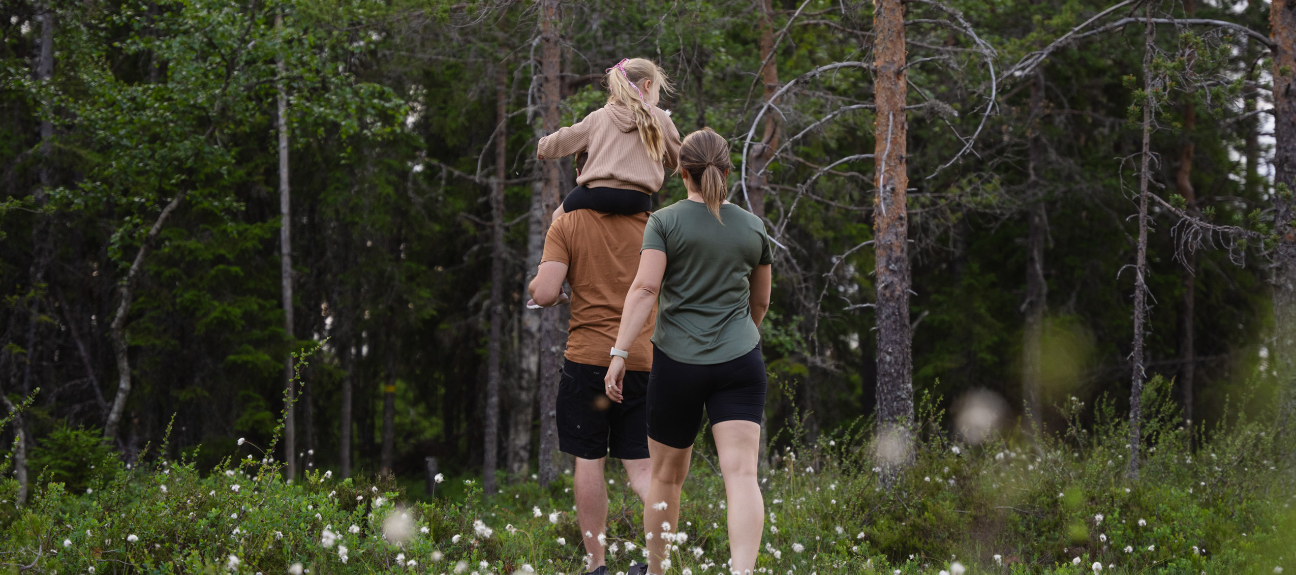 En familj promenerar längs en spång genom en somrig skog. En man bär ett barn på axlarna och en kvinna går bredvid, med ryggen mot kameran.