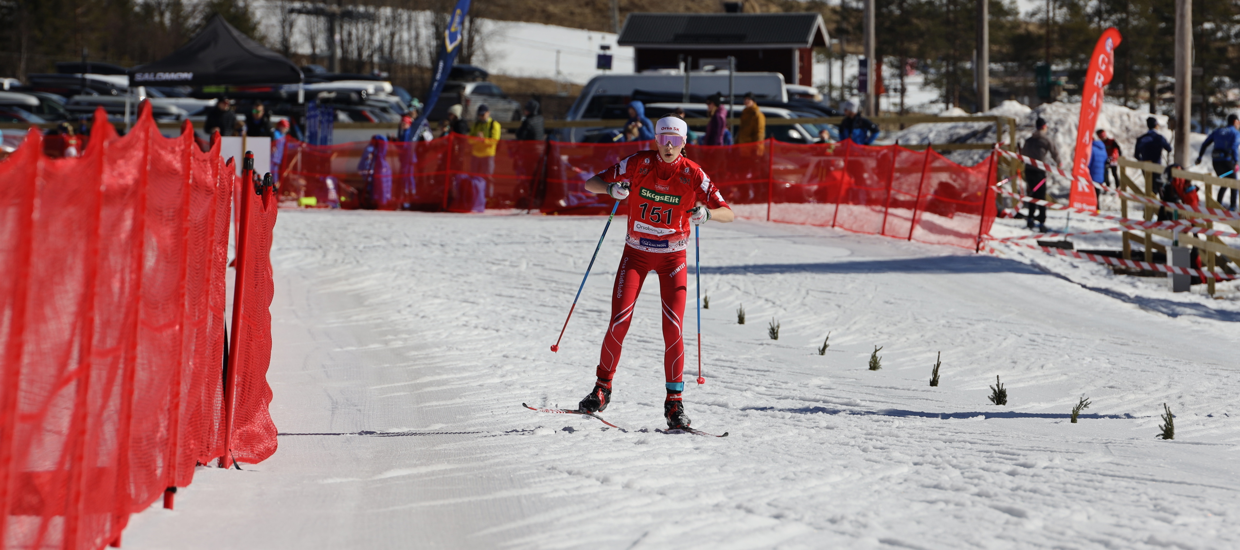 Längdskidåkare med nummerlapp åker i målområde avgränsat med röda nät.
