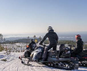 Tre personer på snöskotrar stannar till vid en utsiktsplats med vy över ett vinterlandskap och sjöar i fjärran