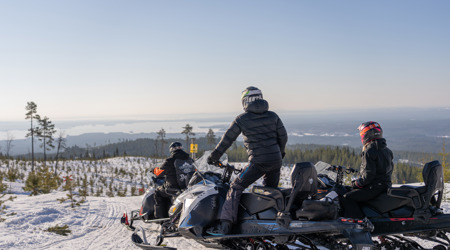 Tre personer på snöskotrar stannar till vid en utsiktsplats med vy över ett vinterlandskap och sjöar i fjärran
