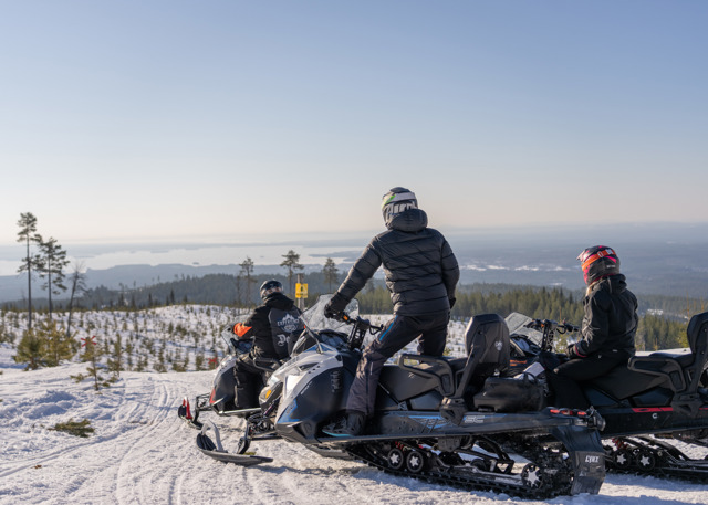 Tre personer på snescootere holder pause ved et udsigtspunkt med udsigt over et vinterlandskab og søer i det fjerne.
