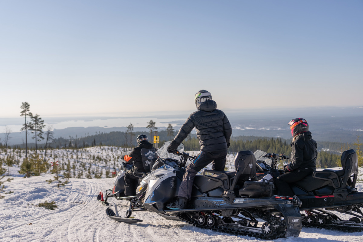 Tre personer på snöskotrar stannar till vid en utsiktsplats med vy över ett vinterlandskap och sjöar i fjärran