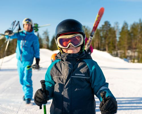 Barn i skitøj med hjelm og skibriller, klar til skiløb i solrigt vinterlandskab.