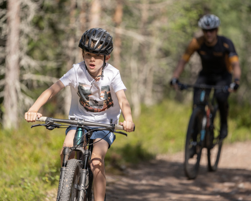 Barn cyklar på skogsstig, vuxen cyklist följer bakom