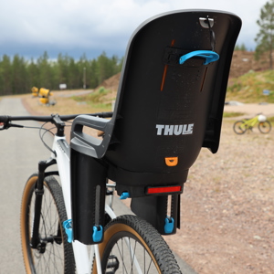 Cykel med barnsits monterad bak, på asfalterad väg i ett skogsområde.