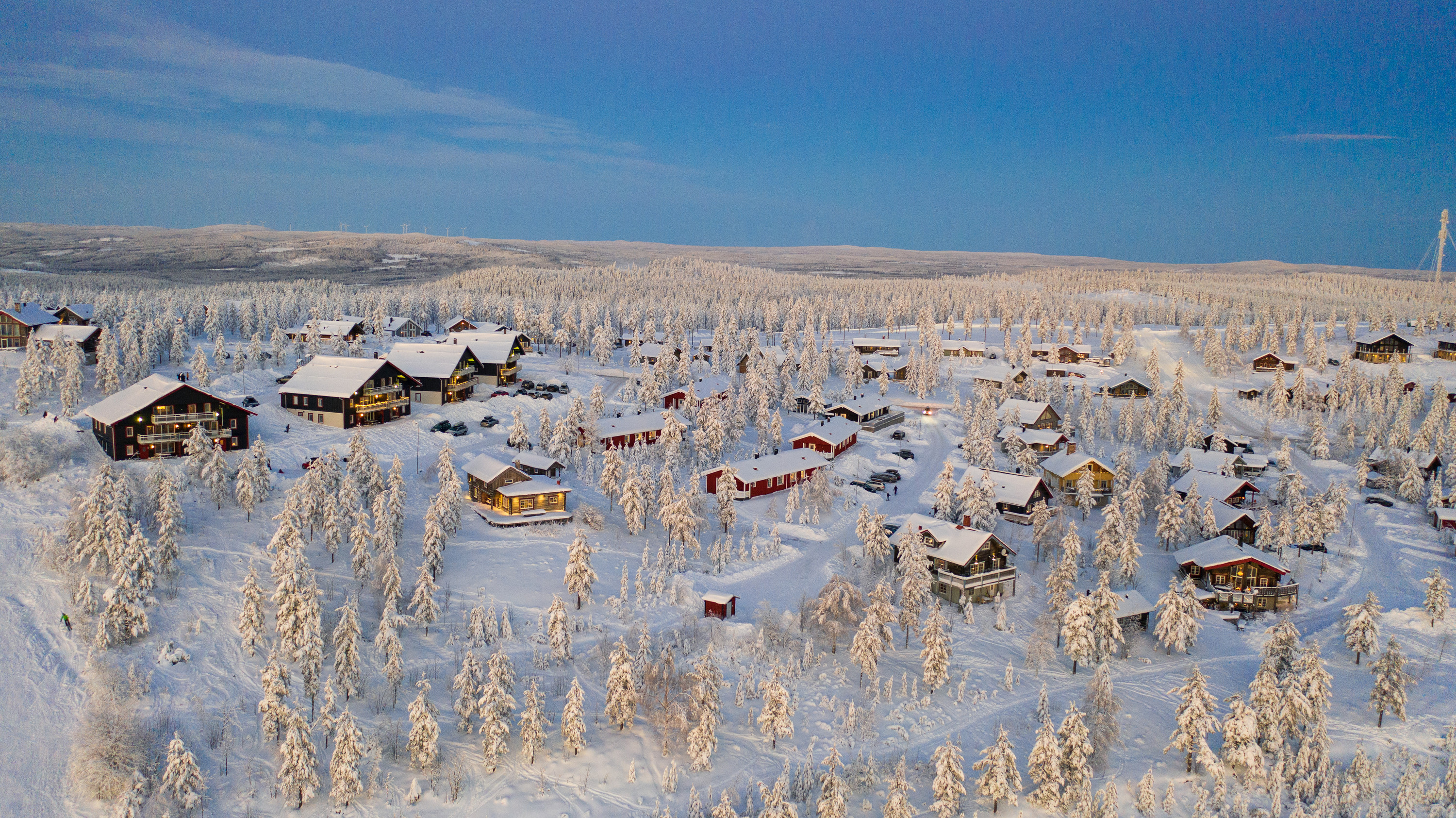 Aerial view of a snow-covered cabin area surrounded by frost-covered trees and blue sky in Orsa Grönklitt.