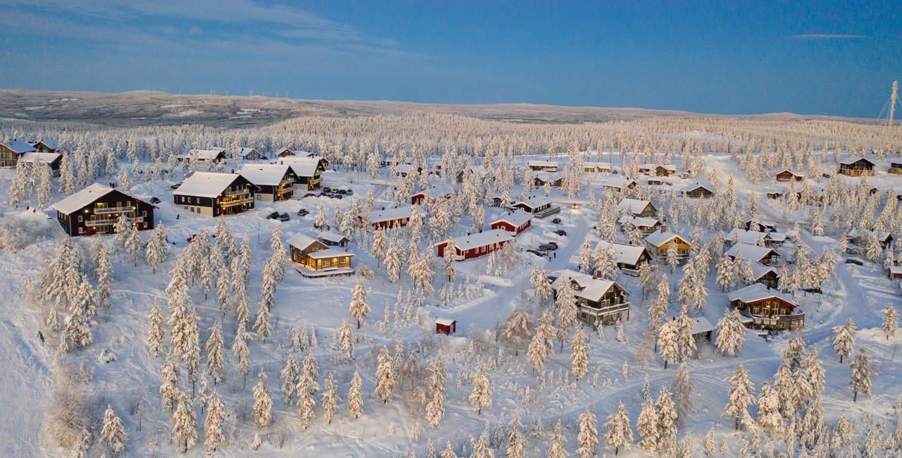 Aerial view of a snow-covered cabin area surrounded by frost-covered trees and blue sky in Orsa Grönklitt.