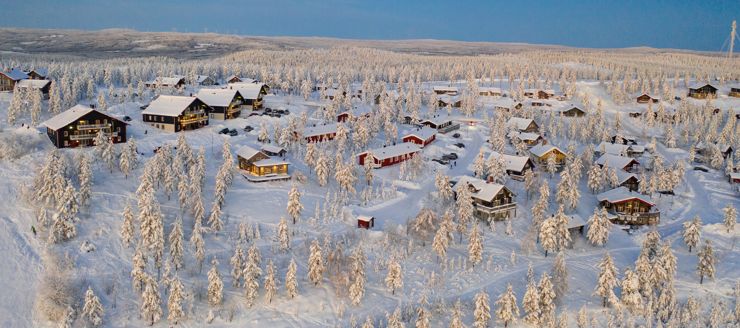Drönarbild över ett snötäckt stugområde omgivet av frostklädda träd och blå himmel i Orsa Grönklitt.