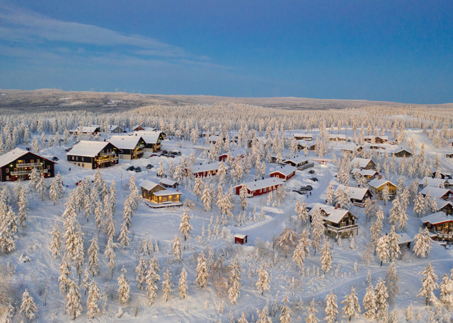 Drönarbild över ett snötäckt stugområde omgivet av frostklädda träd och blå himmel i Orsa Grönklitt.