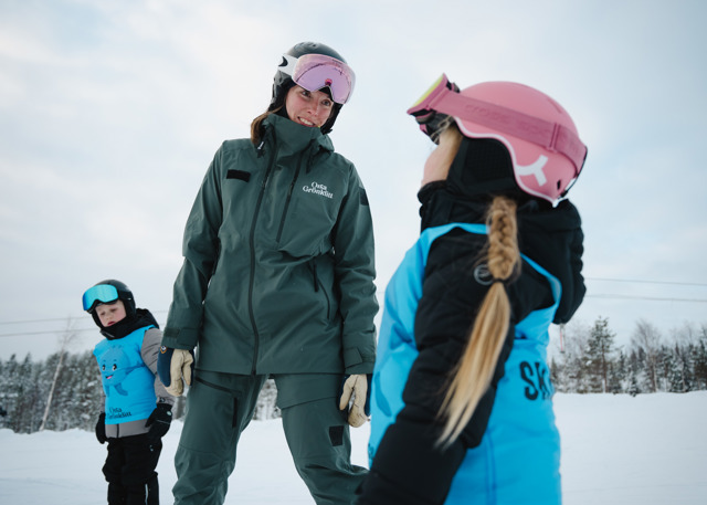 Skidinstruktör hjälper barn under skidlektion i en snötäckt skidbacke.