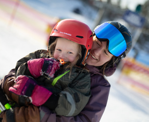 Barn med röd hjälm och stora rosa vantar skrattar medan en vuxen står bakom och kramar om, båda med skidutrustning i solen.