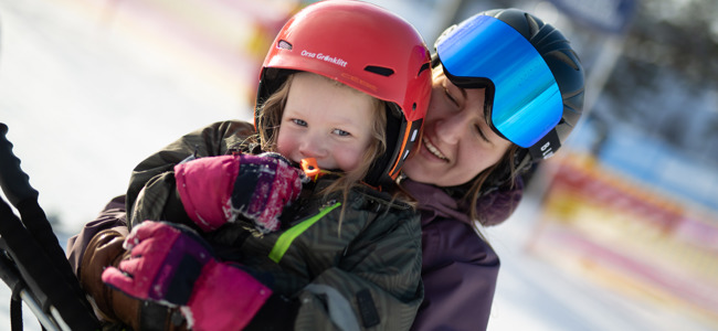 Barn med röd hjälm och stora rosa vantar skrattar medan en vuxen står bakom och kramar om, båda med skidutrustning i solen.