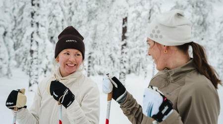 Två personer står på längdskidor och ler mot varandra i en snötäckt skog.