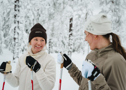 Två personer står på längdskidor och ler mot varandra i en snötäckt skog.