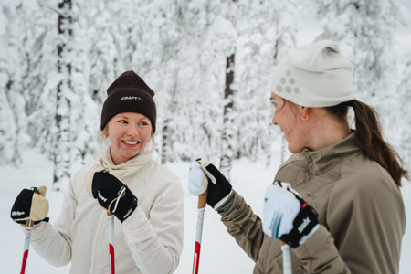Two people on cross-country skis smiling at each other in a snow-covered forest.