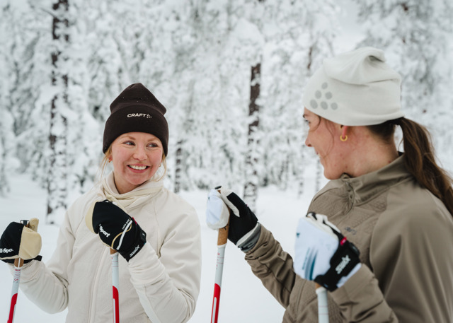 Två personer står på längdskidor och ler mot varandra i en snötäckt skog.