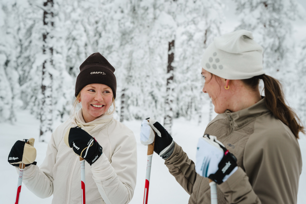 Två personer står på längdskidor och ler mot varandra i en snötäckt skog.