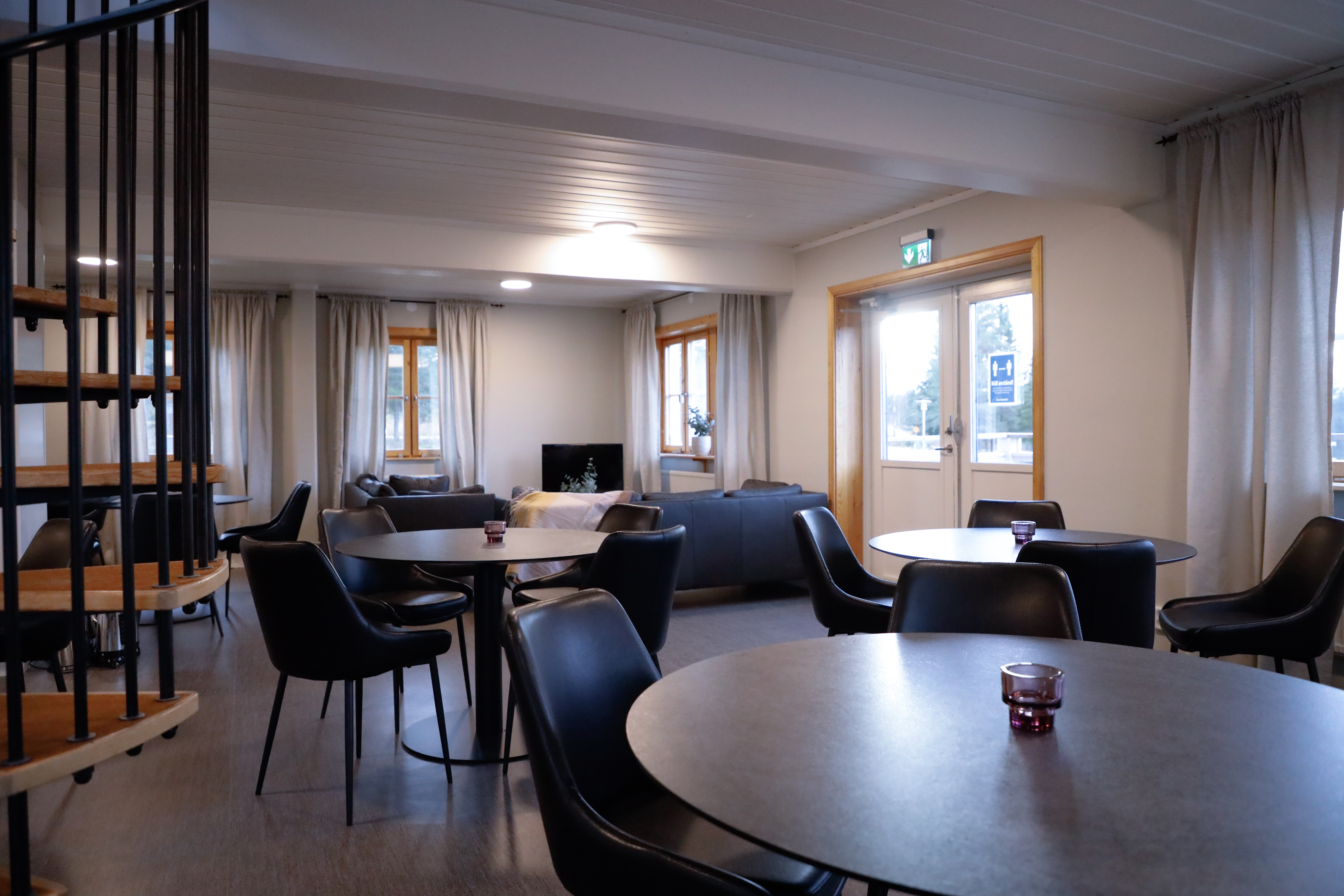 Gemensamt rum med runda bord, svarta stolar, spiraltrappa, soffor och TV.