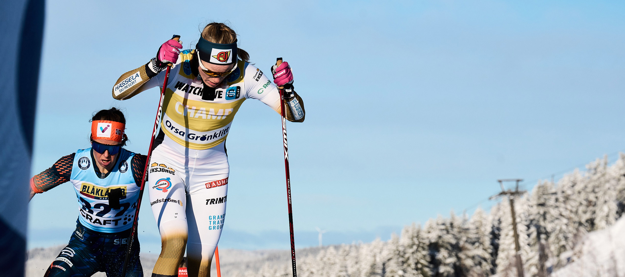 Två längdskidåkare tävlar i en uppförsbacke i vinterlandskap, med snötäckta träd och klarblå himmel i bakgrunden.