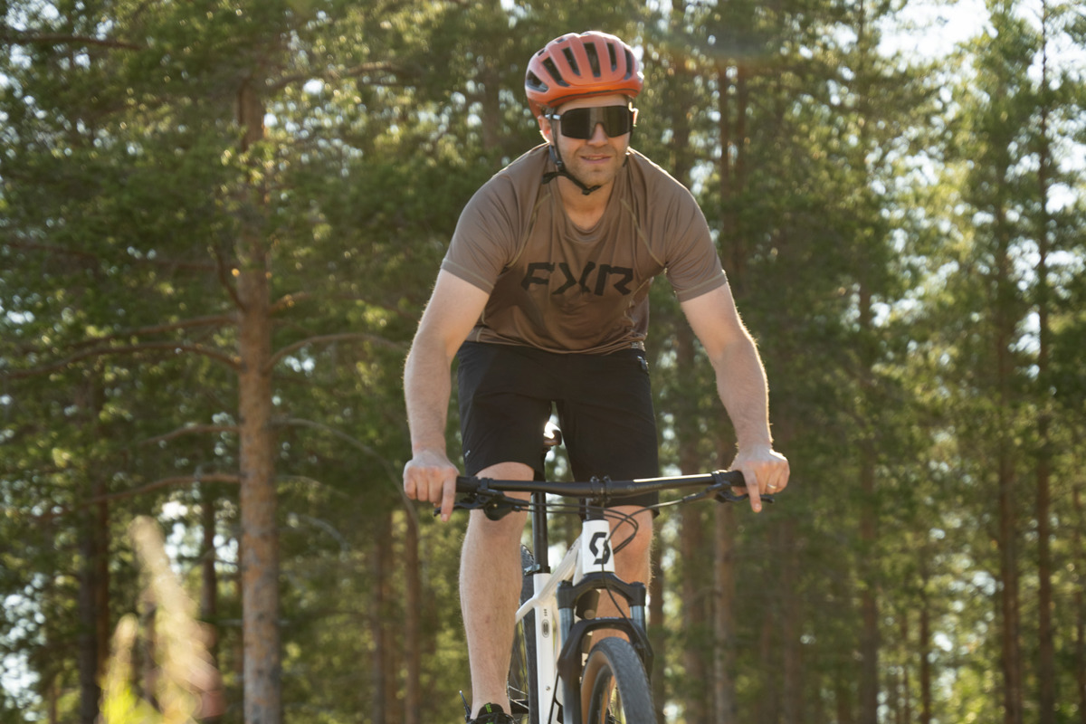 Man med hjälm cyklar på grusväg genom solbelyst tallskog.