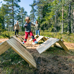 Child balances on wooden obstacles while adults watch in the forest.