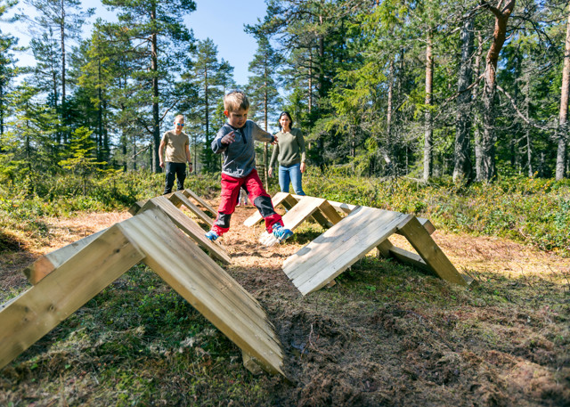 Child balances on wooden obstacles while adults watch in the forest.