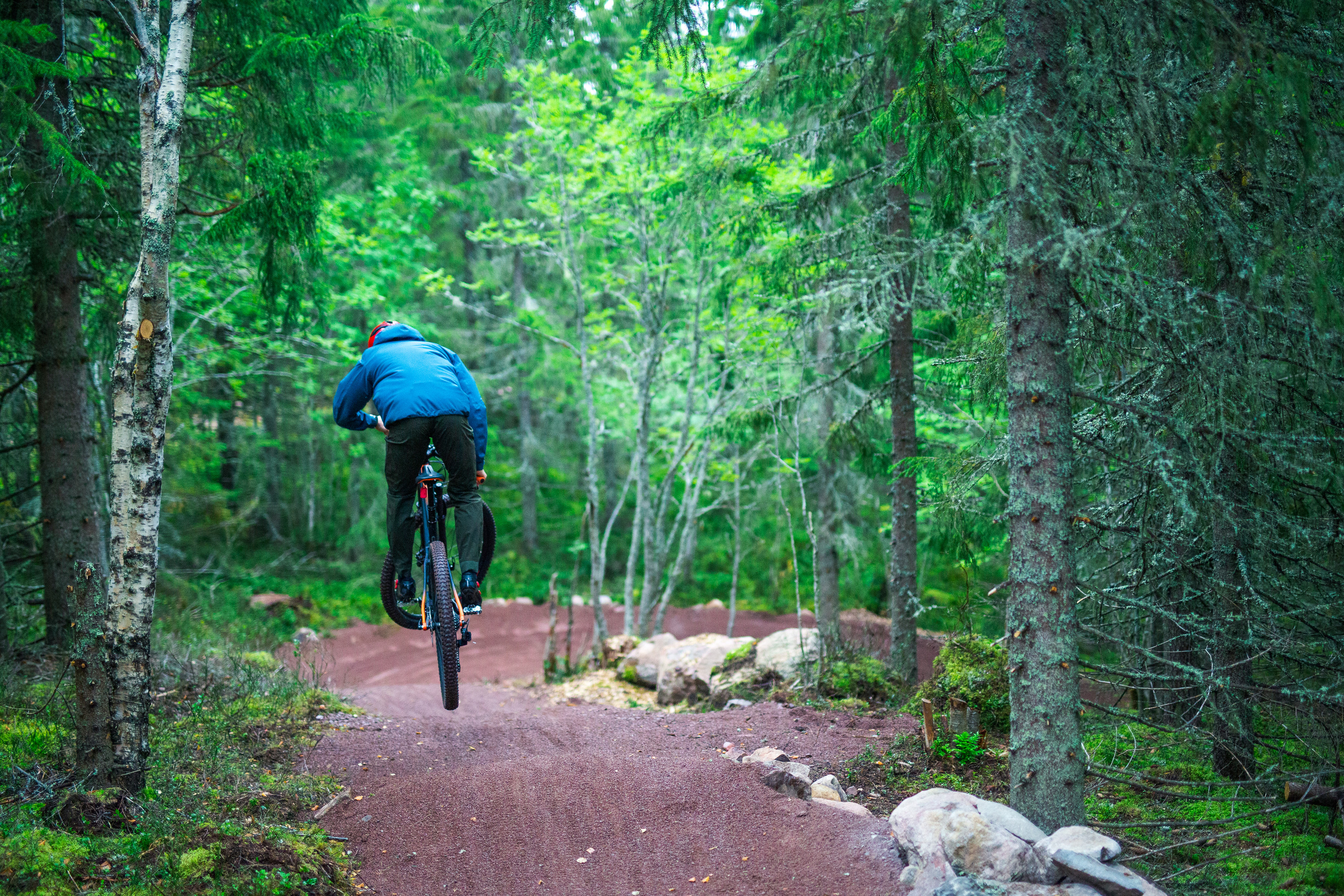 Cyklist i blå jacka som hoppar med mountainbike på skogsstig omgiven av grönskande träd.