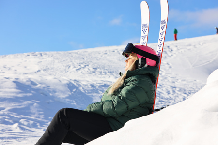 Kvinna med rosa hjälm och skidglasögon sitter lutad mot sina skidor i en solig skidbacke och njuter av vintersolen.