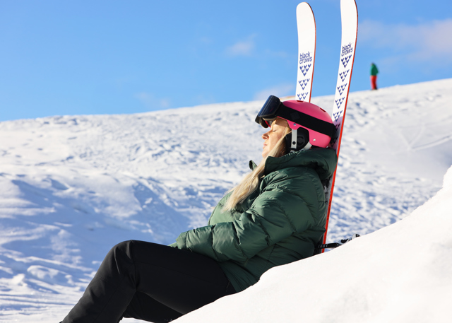 Woman wearing a pink helmet and ski goggles rests against her skis in a sunny slope, enjoying the winter sun.