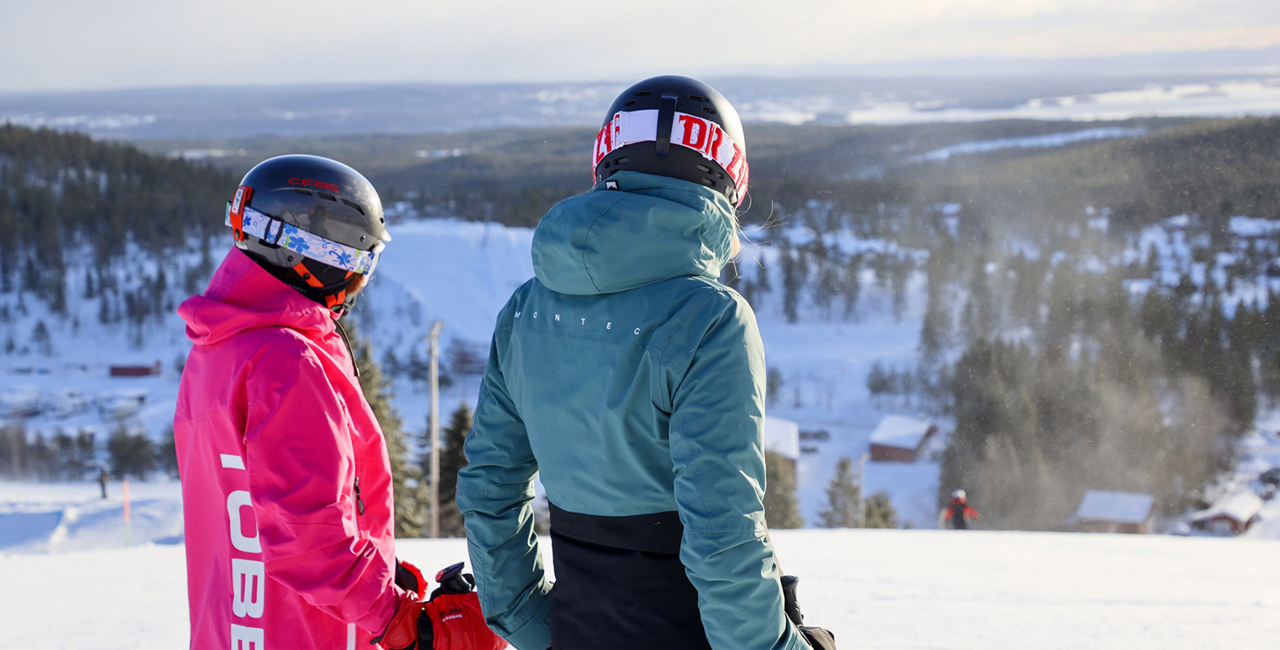 Two skiers wearing helmets look out over a winter landscape from the top of a slope.