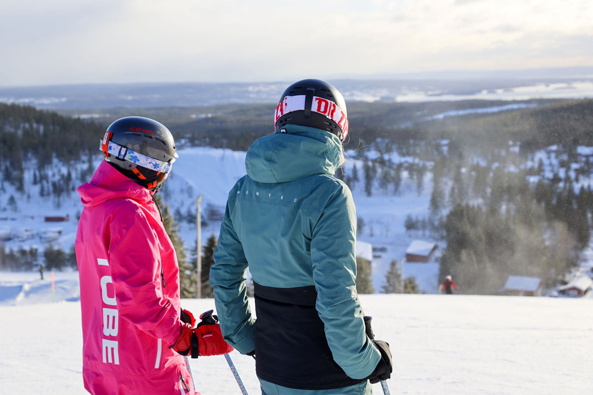 Two skiers wearing helmets look out over a winter landscape from the top of a slope.