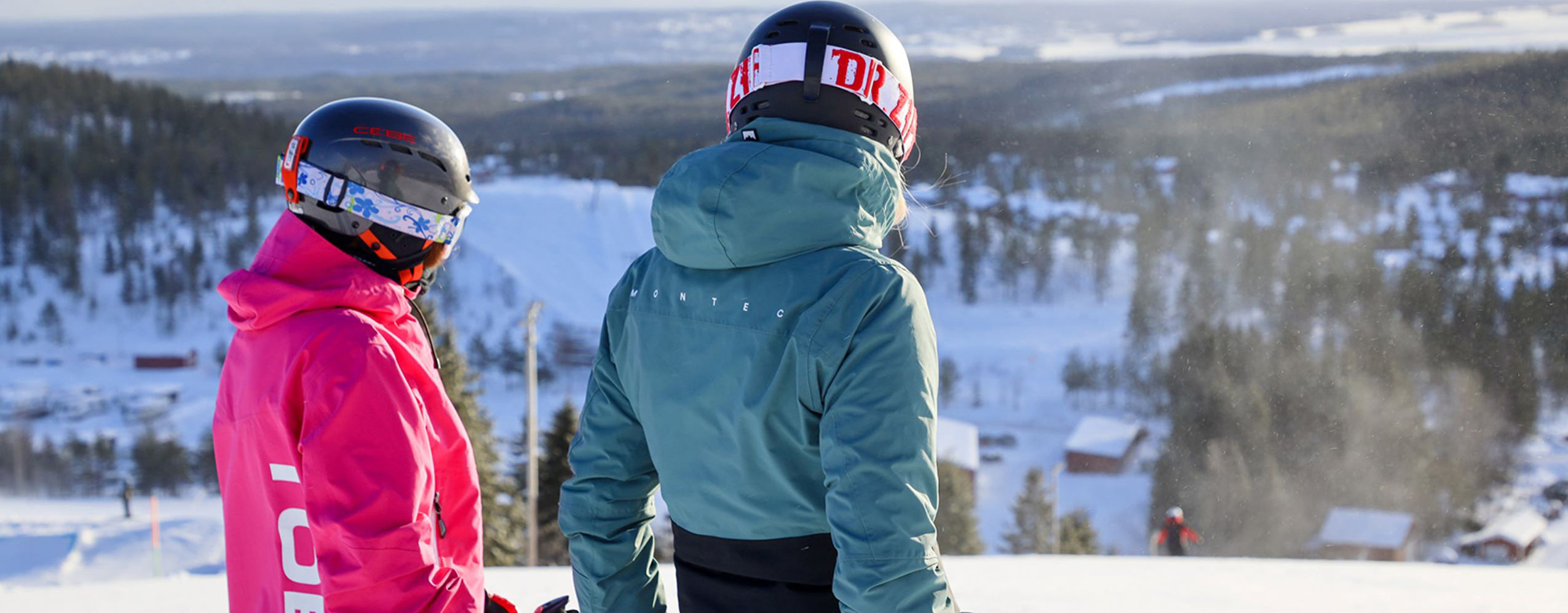 Två skidåkare med hjälm blickar ut över ett vinterlandskap från toppen av en backe.
