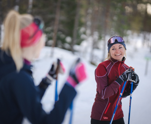 Två kvinnor på längdskidor stannar till i spåret och pratar med varandra en vinterdag i skogen.