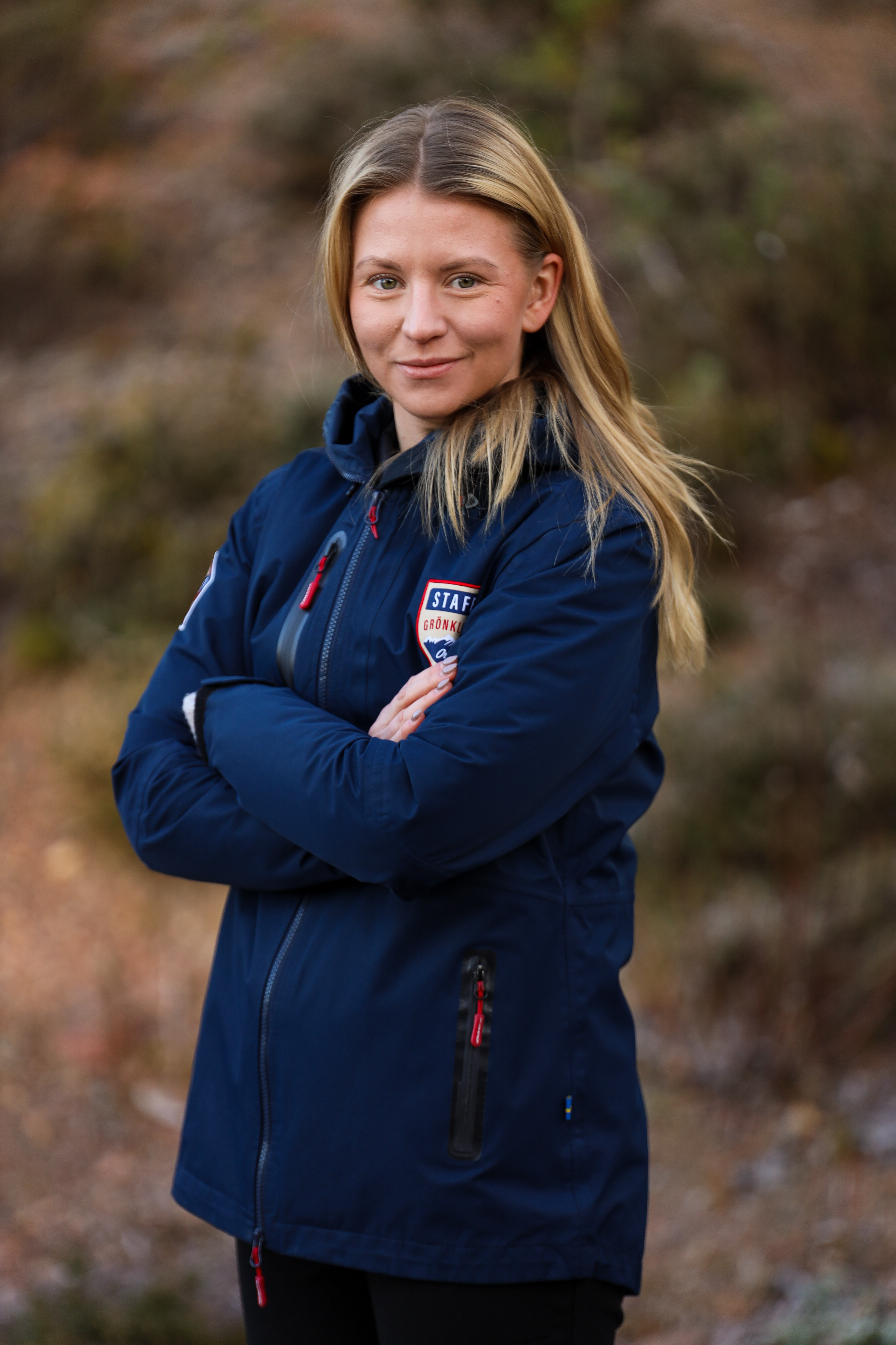 Kvinna med långt blont hår, klädd i blå jacka med Orsa Grönklitt-emblem, står utomhus med armarna i kors och ler mot kameran.