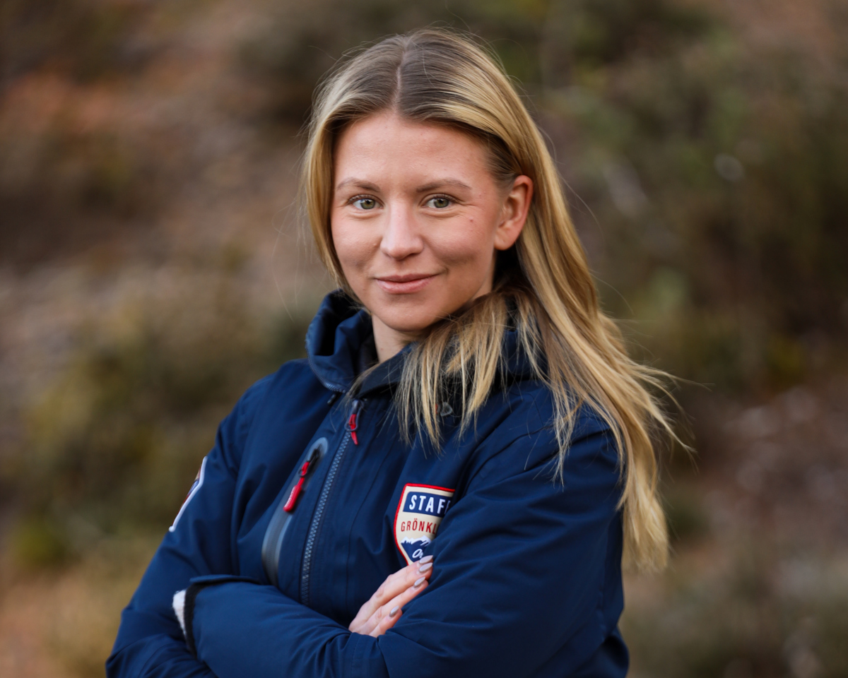 Kvinna med långt blont hår, klädd i blå jacka med Orsa Grönklitt-emblem, står utomhus med armarna i kors och ler mot kameran.