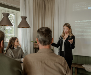En kvinna står vid en projektorduk och håller en presentation för tre personer som sitter runt ett bord i ett mötesrum.