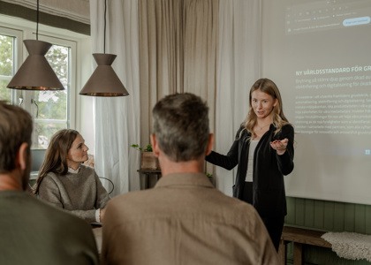 En kvinna står vid en projektorduk och håller en presentation för tre personer som sitter runt ett bord i ett mötesrum.