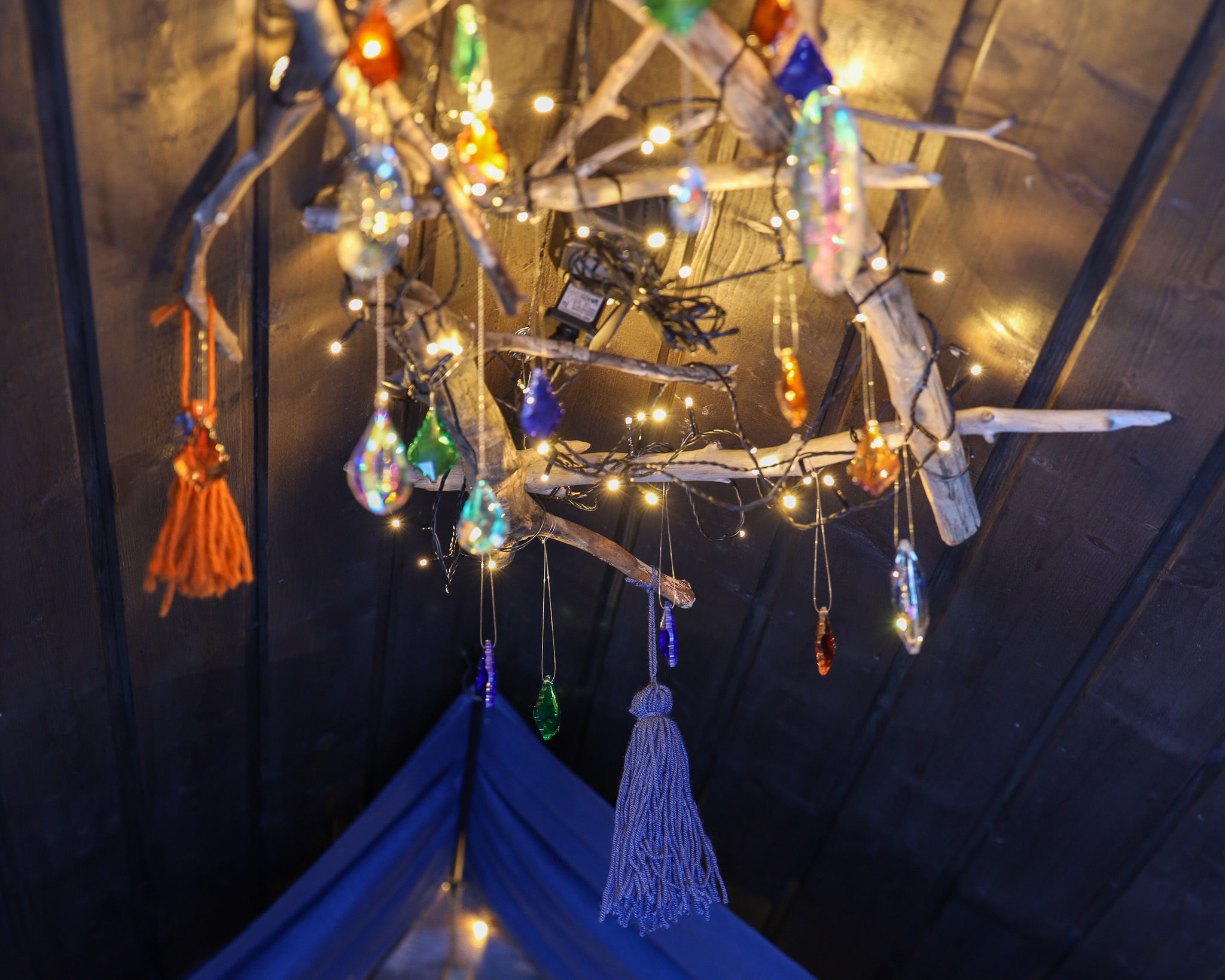 Dekorativ ljuskrona av drivved med hängande glaspärlor och små ljusslingor i varma färger mot ett mörkt träinnertak.